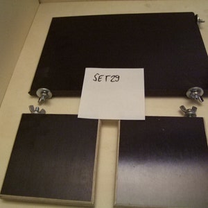 May include: Three black wooden squares with silver metal knobs on each corner. The squares are on a light wood surface. A white piece of paper with the text "SET29" is in the center of the image.