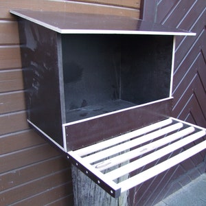 Könnte beinhalten: Ein dunkelbraunes Vogelhaus aus Holz mit weißem Rand und einer weißen Landeplattform. Das Vogelhaus ist an einer Holzwand montiert.