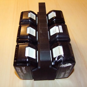 Könnte beinhalten: Ein schwarzer Batteriehalter aus Kunststoff mit sechs Steckplätzen zum Aufbewahren von Batterien. Der Halter hat einen schwarzen Griff oben. Die Batterien sind schwarz und haben weiße Etiketten mit der Aufschrift "Milwaukee 18V Li-Ion".
