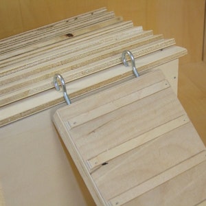 Op de afbeelding: Een houten helling met een scharnierende bovenkant, bevestigd aan een houten doos met metalen haken. De helling is gemaakt van multiplex en heeft een natuurlijke houten afwerking.