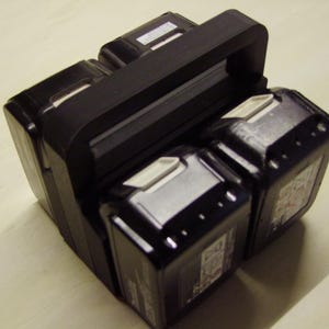 Könnte beinhalten: Ein schwarzer Batteriehalter mit Griff, der vier schwarze Elektrowerkzeugbatterien hält. Die Batterien haben weiße Akzente und Etiketten. Der Halter ist für den Transport mehrerer Batterien konzipiert.