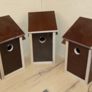 Könnte beinhalten: Drei braune Vogelhäuschen aus Holz mit weißen Verzierungen und runden Eingangsöffnungen. Die Vogelhäuschen sind in Dreiecksform auf einer hellbraunen Oberfläche angeordnet.