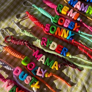 May include: A collection of colourful paracord keychains, each featuring a silver metal lobster clasp and a series of plastic alphabet beads spelling out names like Remi, Bobby, Sonny, Ryder, Mama, Dolly, Dusty, and Maya in various bright colours.
