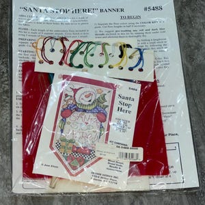 May include: A cross-stitch kit for a Christmas banner with the text "Santa Stop Here". The kit includes floss, aida cloth, and instructions. The finished banner measures approximately 5 inches by 10 inches.