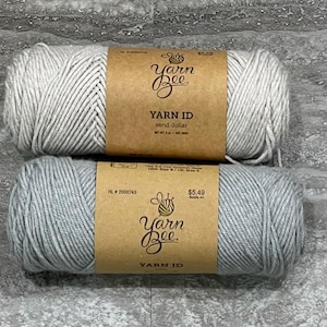 May include: Two skeins of Yarn Bee Yarn ID yarn stacked vertically. The top skein is a light beige color labeled sand dollar, and the bottom skein is a light gray color. Each skein features a brown paper label with the brand logo and text.