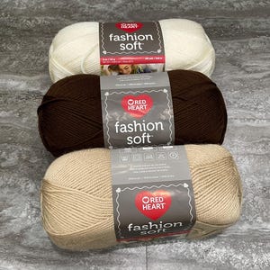May include: Three skeins of Red Heart Fashion Soft yarn stacked vertically. The top skein is off-white, the middle is dark brown, and the bottom is light beige. Each skein features a central paper label with the brand logo and product name.