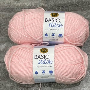 May include: Two balls of light pink Basic Stitch Premium yarn. Each ball has a label with the brand name and icons for knitting projects. The yarn is soft and fluffy, perfect for various knitting and crochet projects.