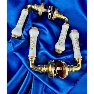 Peut inclure: Deux ensembles de poignées de porte avec des prises en céramique blanche ornées d'un motif floral rose délicat et de feuilles vertes. Les poignées présentent une quincaillerie en métal doré poli, des cols incurvés et des rosaces circulaires.