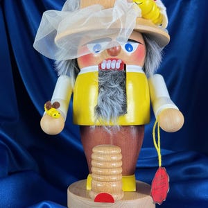 May include: Wooden nutcracker figurine of a beekeeper, wearing a yellow jacket, brown pants, and a hat with a bee. The figurine has a white beard, blue eyes, and a red mouth. It holds a bee on one hand and a beehive on the base.