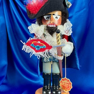 May include: A wooden nutcracker depicting Porthos from The Three Musketeers. The figure wears a black hat with a red feather, a white lace collar, and a blue cape. It holds a sword and a shield with a red and blue design. The base is wooden.