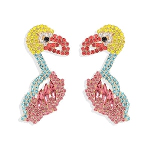 Puede incluir: Un par de pendientes con forma de flamenco. Los pendientes están adornados con pedrería de colores en tonos amarillos, rojos, rosas y azules. Las cabezas de los flamencos son amarillas con picos rojos y ojos negros. Los cuerpos son rosas con detalles azules.