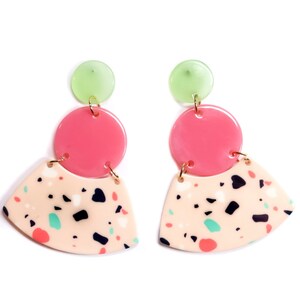 May include: A pair of colorful earrings with a geometric design. Each earring features a light green circle at the top, a pink circle in the middle, and a beige, fan-shaped bottom with black, blue, and pink speckles. Gold-colored metal connects the pieces.