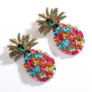 Könnte beinhalten: Ein Paar goldfarbene Ohrringe in Form von Ananas. Die Ohrringe haben ein detailliertes Design mit grünen, blauen, rosa, gelben und orangefarbenen Edelsteinen. Die Ananas-Oberteile sind grün, der Körper ein Mosaik aus bunten Edelsteinen.