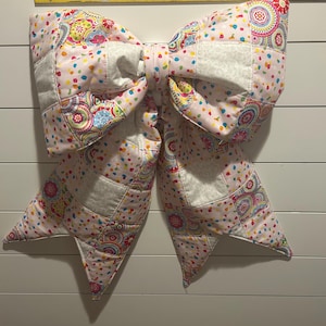 May include: A large, quilted bow in shades of pink, white, and multicolored patterns. The bow is made of patchwork squares and features a confetti-like dot pattern. The bow measures approximately 19 inches across.