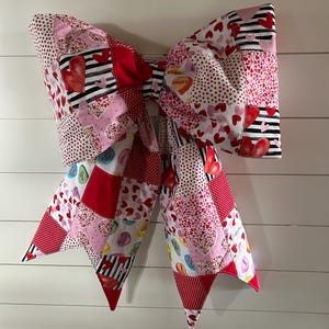 May include: A large, quilted bow in shades of red, pink, and white, featuring heart and stripe patterns. The bow is made of fabric squares and is likely a decorative item for Valentine's Day or a similar occasion.