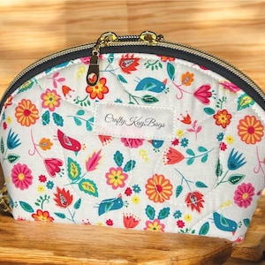 May include: A quilted, white cosmetic bag with a colorful floral and bird pattern. The bag features a gold zipper and the text "Crafty Kay Bags" on a small label. The bag is on a wooden surface.
