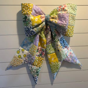 May include: A large, colorful patchwork bow with a variety of floral patterns in yellow, blue, green, and purple. The bow is made of quilted fabric and has a metal hook for hanging. The bow is a decorative item.