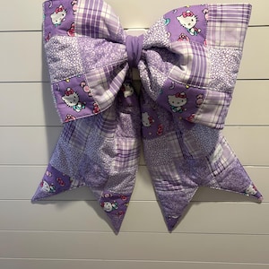 May include: A large, quilted bow in shades of purple, featuring a patchwork design with various patterns, including Hello Kitty motifs. The bow is tied in the center and hangs against a white, horizontal-slat background.