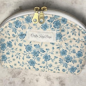 May include: A quilted, half-moon shaped cosmetic bag with a floral pattern in light blue and cream. The bag has a white zipper and two gold-colored zipper pulls. The label on the bag reads "Crafty Kay Bags".