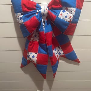 May include: A large, quilted bow in red, white, and blue. The bow is made of fabric squares, with a star pattern on some squares. The bow is hanging on a white wall. The bow is a decorative item.