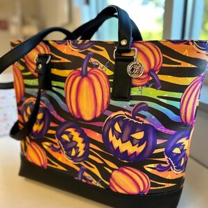 May include: A Halloween-themed tote bag with a black base and handles. The bag features a vibrant pattern of orange pumpkins, purple jack-o'-lanterns, and colorful stripes against a black zebra-stripe background. A silver charm hangs from the handle.