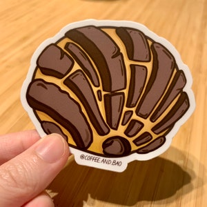Op de afbeelding: Een bruine en gele sticker met een cartoonillustratie van een concha, een Mexicaans zoet brood. De sticker heeft de tekst "@COFFEE.AND.BAO" onderaan.