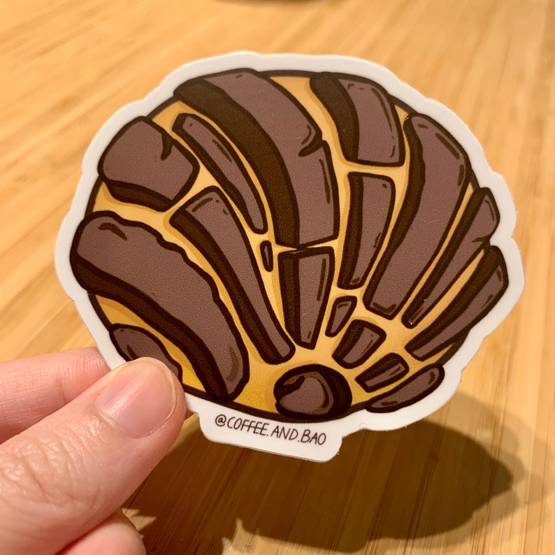 Op de afbeelding: Een bruine en gele sticker met een cartoonillustratie van een concha, een Mexicaans zoet brood. De sticker heeft de tekst "@COFFEE.AND.BAO" onderaan.