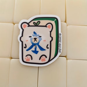 May include: A white bear sticker with a blue and white design on a green background. The sticker is shaped like a mahjong tile and has the text "@COFFEE_AND_BAO" on the side.