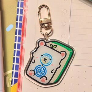 May include: A white bear with a blue and white wheel design is sleeping on a green and white rectangle. The bear is attached to a gold key chain.