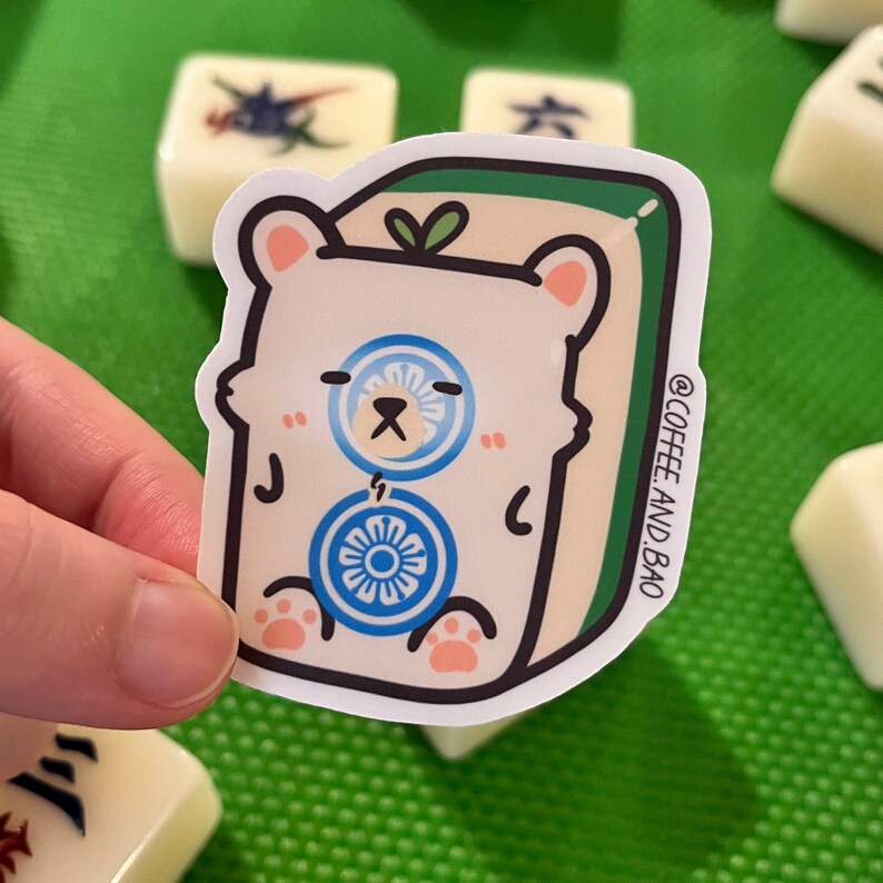 May include: A white bear sticker with blue and white circular designs, sitting on a green and white mahjong tile. The sticker has a green leaf on top and the text "@COFFEE_AND_BAO" on the side.