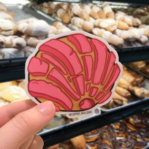Op de afbeelding: Een roze en bruine concha, een soort Mexicaans zoet brood, wordt in een hand gehouden. Het brood is versierd met een swirl-patroon. De tekst "@COFFEE AND BAO" is zichtbaar onderaan de afbeelding.
