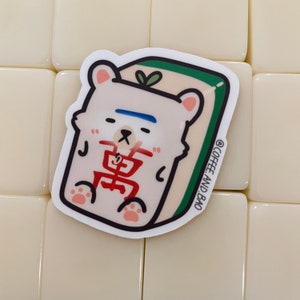 May include: A white bear sticker with a blue stripe across its forehead, sitting on a green and white mahjong tile. The tile has the Chinese character "萬" in red. The sticker has the text "@COFFEE_AND_BAO" in the bottom right corner.