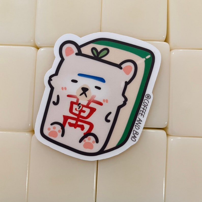 May include: A white bear sticker with a blue stripe across its forehead, sitting on a green and white mahjong tile. The tile has the Chinese character "萬" in red. The sticker has the text "@COFFEE_AND_BAO" in the bottom right corner.