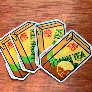 May include: Four holographic stickers of a yellow and brown cartoon box with the text "Lemon Tea" and "維他 Vita" in red and black. The stickers are on a wooden surface.