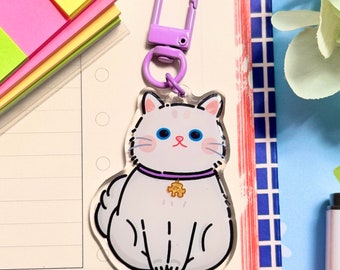 May include: A white cat keychain with purple collar and a gold charm. The keychain is made of clear acrylic and has a purple metal ring.
