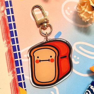 May include: A gold keychain with a clear acrylic charm featuring a cartoon bread slice with a smiling face. The bread is brown with a red crust and has a cute expression.