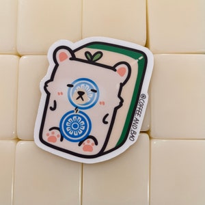 May include: A white sticker with a cartoon bear wearing a blue and white patterned shirt. The bear is sitting on a green and white background. The sticker says "@COFFEE_AND_BAO" in the bottom right corner.