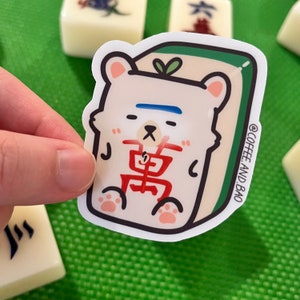 May include: A white sticker of a cartoon bear wearing a red and white outfit with the Chinese character for "East" on its chest. The bear is sitting on a green and white mahjong tile. The sticker has a white border and the text "@COFFEE AND BAO" in the bottom right corner.