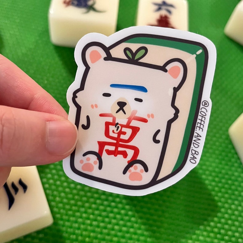 May include: A white sticker of a cartoon bear wearing a red and white outfit with the Chinese character for "East" on its chest. The bear is sitting on a green and white mahjong tile. The sticker has a white border and the text "@COFFEE AND BAO" in the bottom right corner.