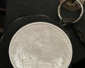 May include: A silver coin featuring a knight in armor holding a chalice, with the word "GRAIL" arched above. The coin is displayed on a clear stand, with a dark background and a metal ring visible.