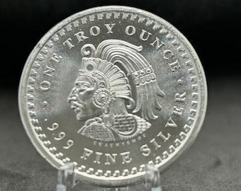 May include: A silver coin with a detailed relief of a figure wearing a headdress. The coin is marked with "ONE TROY OUNCE .999 FINE SILVER". The coin is displayed on a clear stand against a dark background.