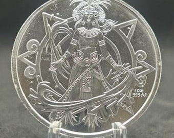 May include: A silver coin featuring a detailed relief of a figure with outstretched arms, adorned with a headdress and intricate clothing. The coin is surrounded by a decorative border with geometric shapes and text. The coin is displayed on a clear stand.