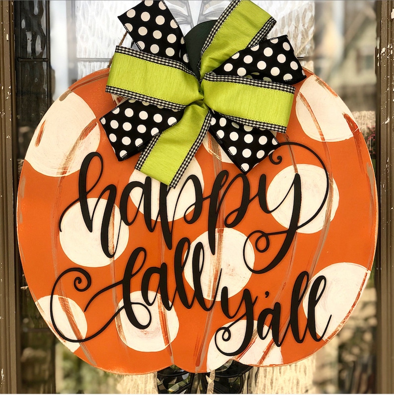 May include: Orange pumpkin-shaped door hanger with white polka dots and the text "happy fall y'all" in black. A black and white polka dot bow with a green ribbon is attached to the top.