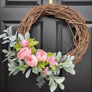 Op de afbeelding: Een roze en groene bloemenkrans met een basis van wijnstokken. De krans is versierd met roze pioenen en eucalyptusbladeren.