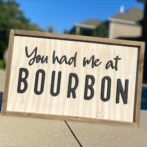 May include: A wooden sign with a brown frame and a white background. The sign says "You had me at BOURBON" in black lettering.