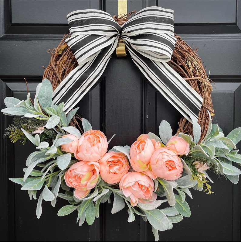 May include: A black and white striped ribbon bow decorates a grapevine wreath with a cluster of peach-colored peonies and dusty miller greenery.