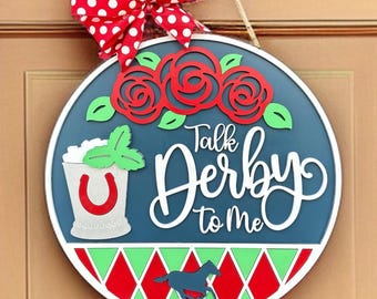 May include: A round wooden door hanger with a blue background and white text that says "Talk Derby to Me". The door hanger is decorated with red roses, green leaves, a mint julep, and a running horse. The door hanger is also decorated with a red and green diamond pattern.