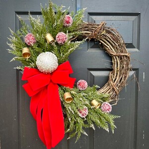 May include: A Christmas wreath with a grapevine base, adorned with evergreen branches, red velvet bow, gold bells, and frosted red and white ornaments. This festive wreath is perfect for holiday decor.