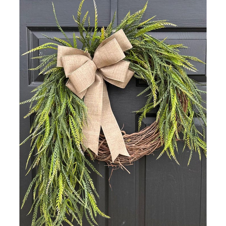 May include: A brown grapevine wreath with a burlap bow. The wreath is decorated with green artificial greenery that hangs down from the wreath.