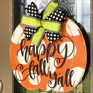 May include: Orange pumpkin-shaped door hanger with white polka dots and the text "happy fall y'all" in black script. A green and black polka dot bow is attached to the top.